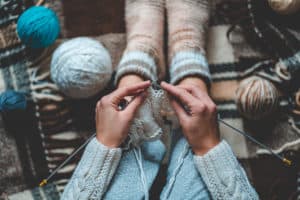 Cozy knitting woman in knitted winter warm socks and in pajamas enjoys knit work on brown checkered plaid blanket at home in cozy winter time. Top view